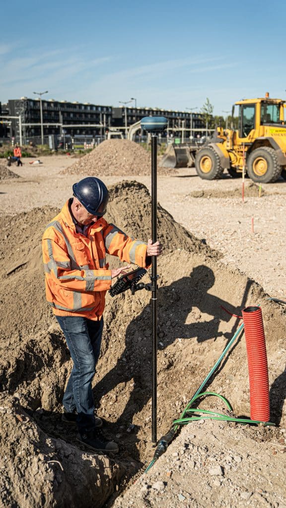 Landmeter in oranje jas staat in een sleuf met kabels en bedient een tablet aan een GNSS-stok, shovel achter