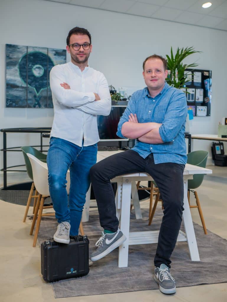 Twee collega's poseren bij een tafel, één zet zijn voet op een zwarte marXact-koffer