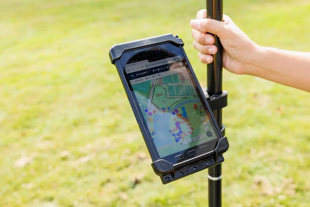 Hand holding a survey pole with a tablet showing a map on a grass field