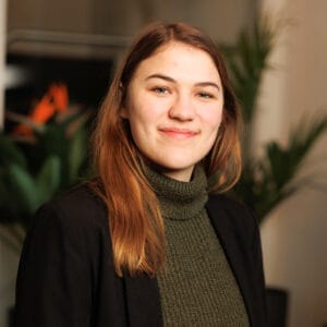 Portret van een lachende vrouw met lang rood-bruin haar in groene coltrui en zwart colbert voor kantoorplanten