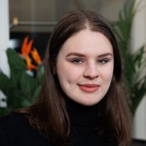 Lachende vrouw met lang bruin haar in een zwarte coltrui, met groene planten op de achtergrond