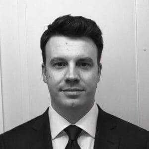 Zwart-witportret van een man met donker haar in pak met stropdas tegen een lichte muur