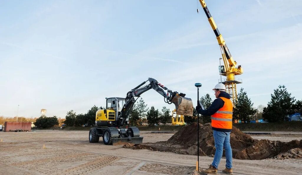 A surveyor with a UNI-GR2 on a pole next to an excavator on a graded road surface