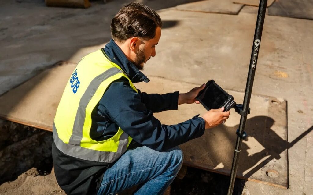 A groundworker kneeling at an open trench in London, taking a point with the UNI-GR2 and a tablet