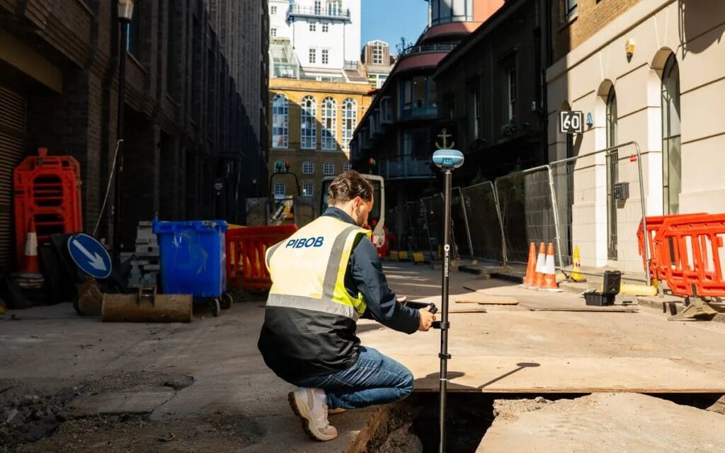 A utilities crew recording an open service trench on a London street behind red barriers