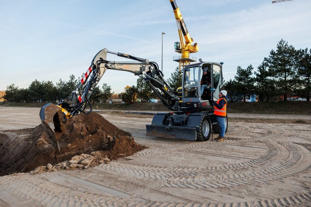 Wheeled excavator with GNSS receivers digging soil, surveyor with pole alongside and a tower crane behind
