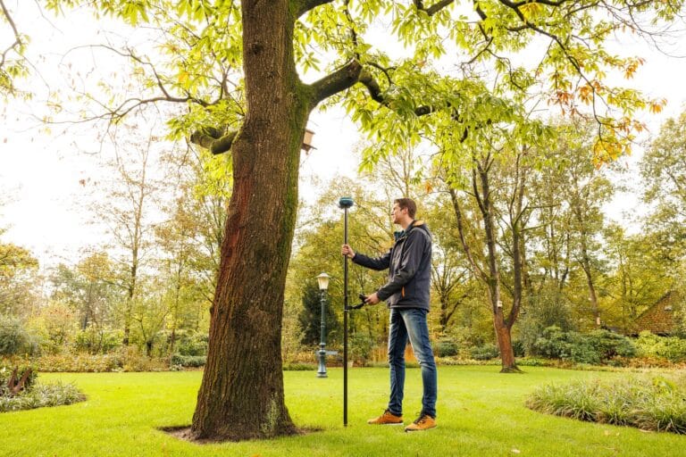 Man holds a pole with a UNI-GR2 receiver next to a tree on a lawn, a lamp post behind