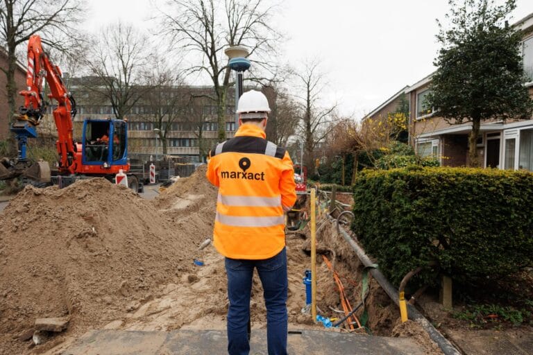 Worker in a marXact jacket seen from behind with survey pole at a trench, red excavator and houses ahead