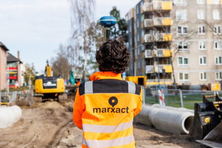 Surveyor in a marXact jacket looking over a works site with pipes and an excavator, UNI-GR2 above him