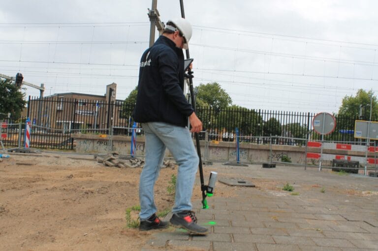Worker in a helmet staking out with a UNI-GR2 pole on a site with a railway and overhead lines behind