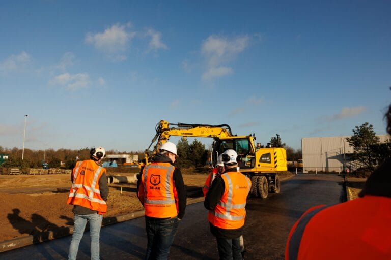Participants in orange vests watch a yellow road-rail excavator on a road