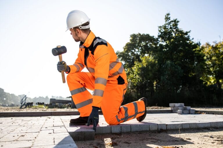 Paver in orange workwear and white hard hat kneels with a mallet on new paving