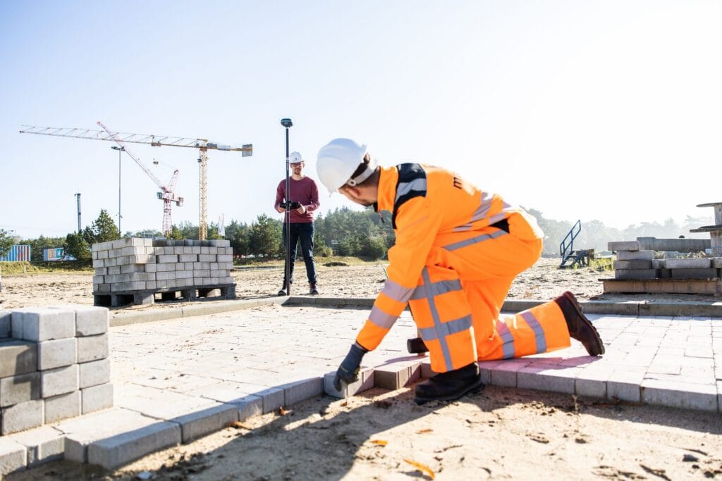 Paver laying kerb blocks with a person holding a UNI-GR2 on a pole in the background