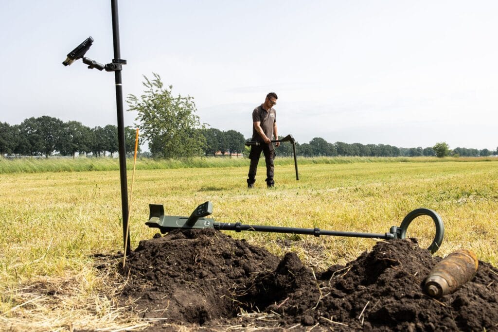Survey pole with tablet holder at a soil mound with shell casing and detector, specialist behind