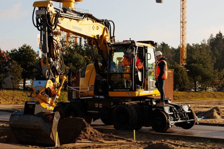 Yellow road-rail excavator with GNSS digs sand in warm light, participant beside it