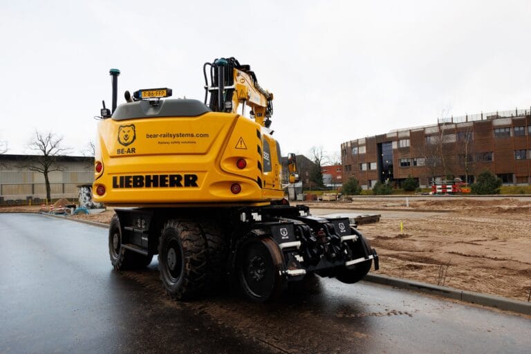 Yellow road-rail wheeled excavator with GNSS receivers on a wet road beside the training site