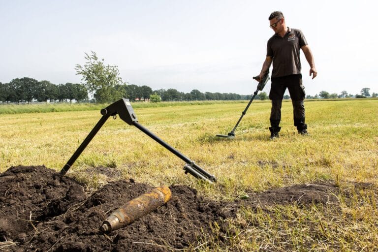 Rusty shell casing on a soil mound in the foreground, specialist with a metal detector behind