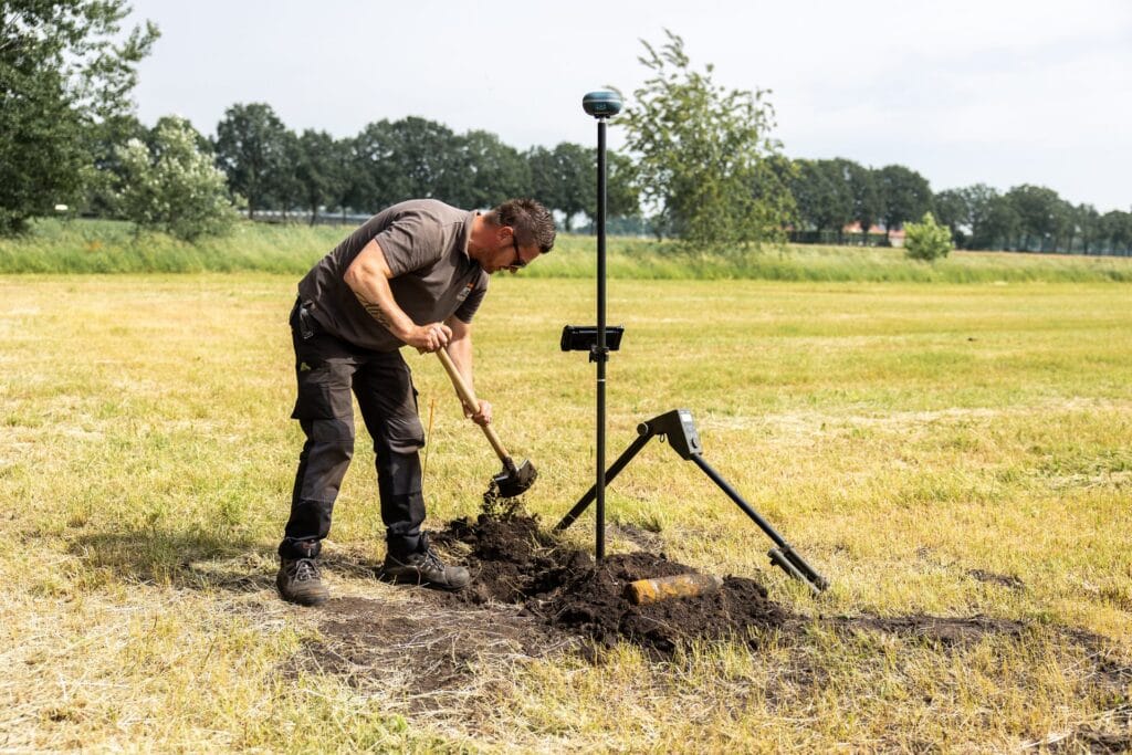 Specialist digging with a spade beside a UNI-GR2 on a pole and a detector in a meadow
