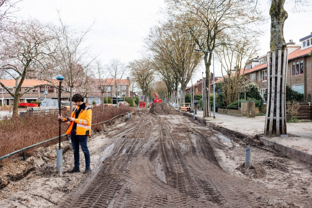 Wide view of a muddy street under construction lined with trees and a surveyor with a UNI-GR2