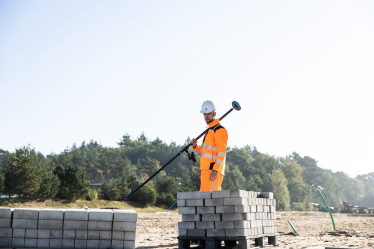 Surveyor in orange workwear carries a UNI-GR2 pole over his shoulder by pallets of blocks