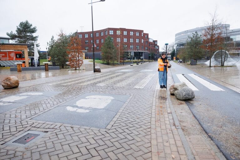 Distant surveyor with an RTK pole on a wet brick-paved street with a cycle path