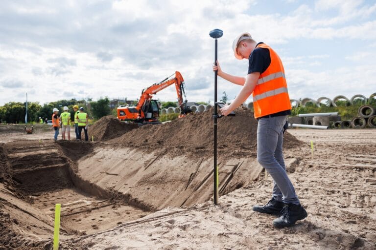 Man in an orange vest places a GNSS pole beside an excavated trench with stakes, excavator in the distance