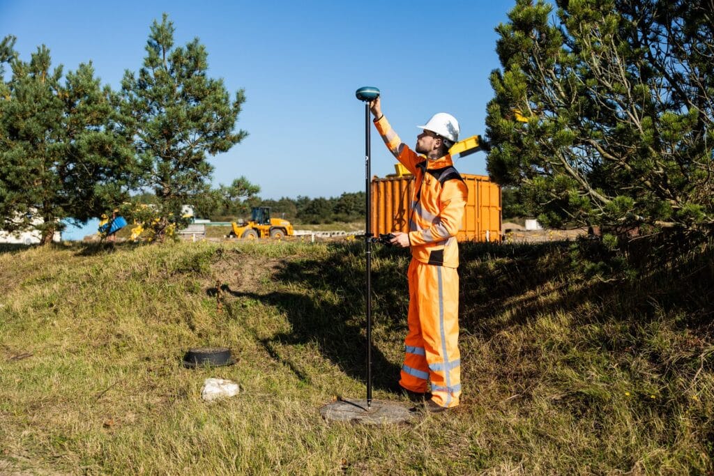 Surveyor in orange workwear reaches up to the UNI-GR2 on his pole among pine trees