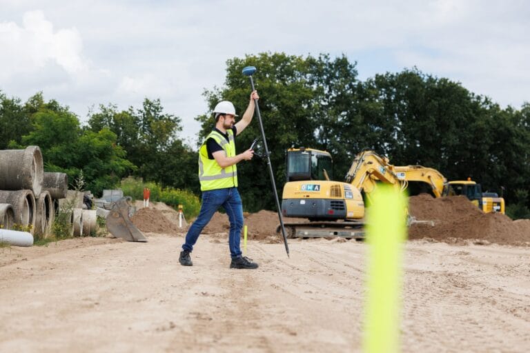 Surveyor in a yellow vest holds a tilted GNSS pole on sandy ground, excavators and pipes behind