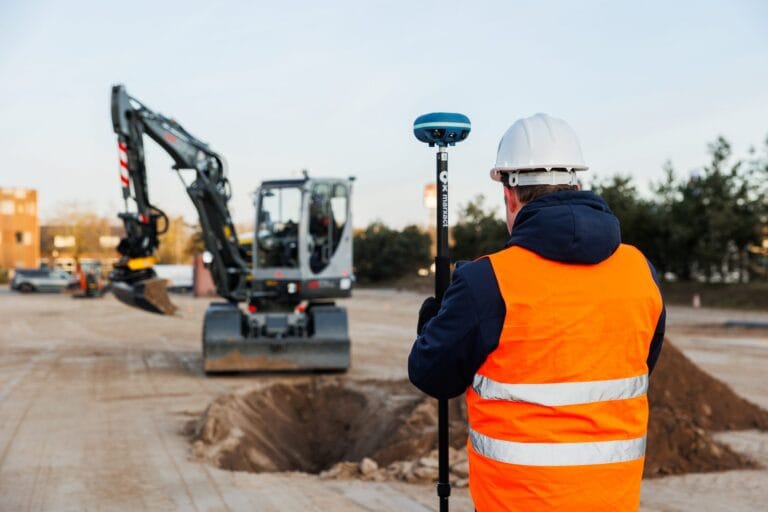 Surveyor in a white hard hat with a UNI-GR2 pole seen from behind, watching an excavator with machine control dig