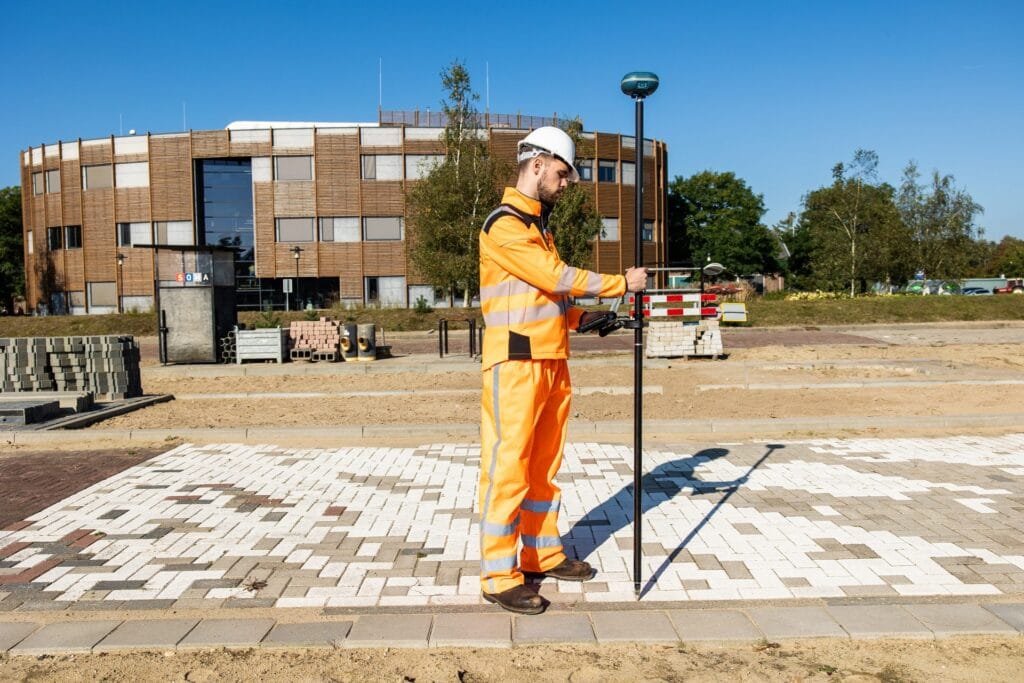 Surveyor in orange workwear with a UNI-GR2 on a pole on newly laid paving in front of a round building