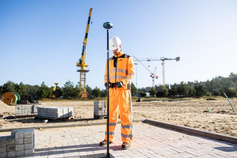 Surveyor in orange workwear with a UNI-GR2 on a pole on a paving site with a crane behind