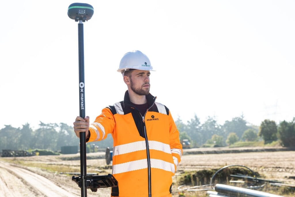Surveyor in an orange marXact jacket and white helmet with a UNI-GR2 on a pole on an open building site