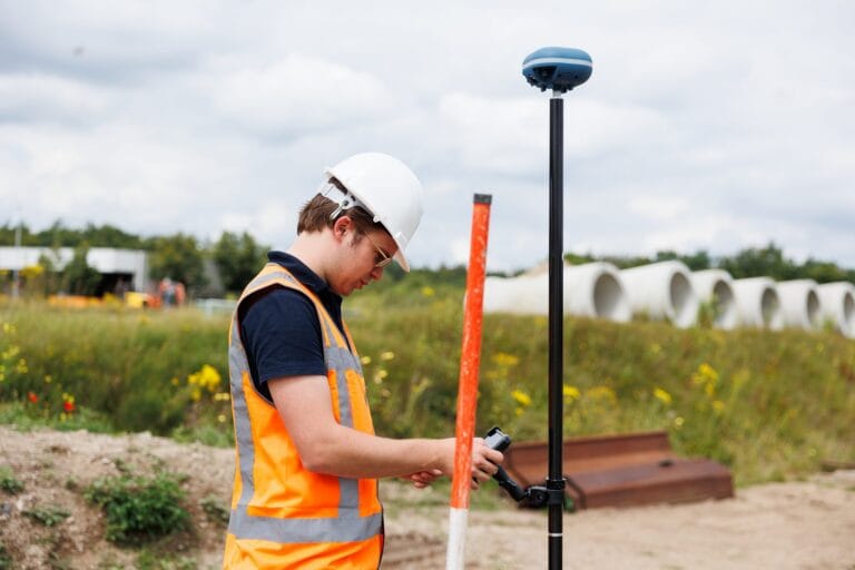 Surveyor in a helmet and orange vest measures with a GNSS receiver on a pole and a tablet