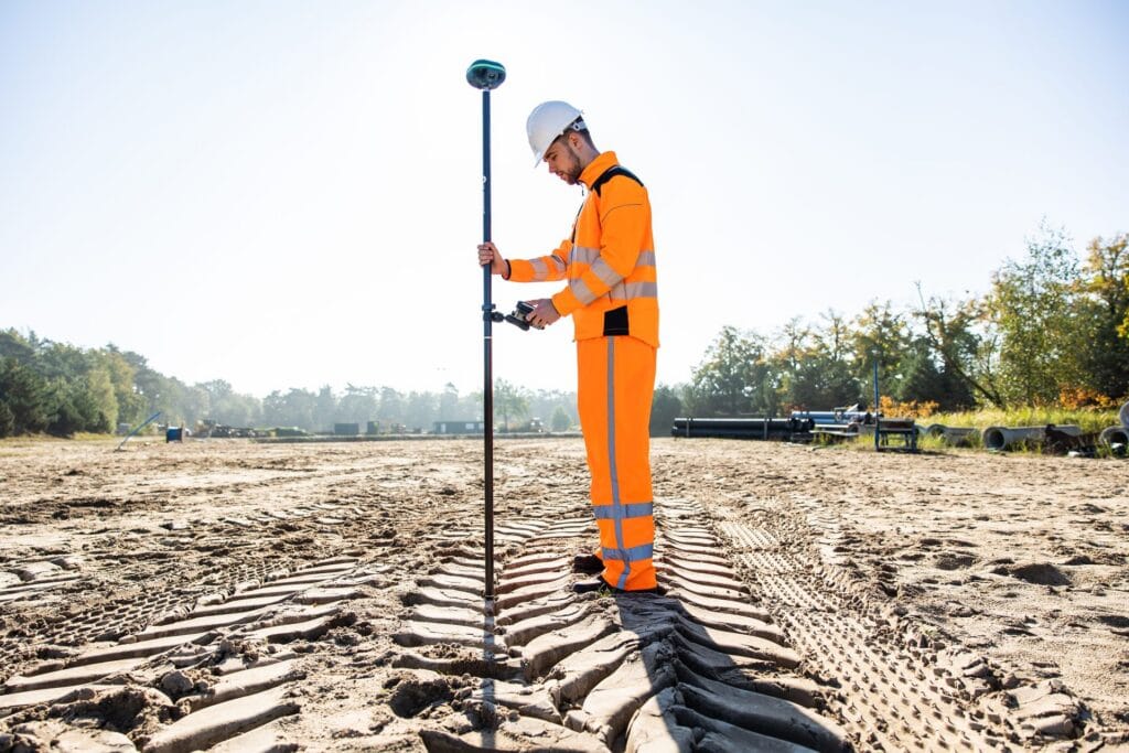 Surveyor in orange workwear with a UNI-GR2 on a pole on sandy ground with tyre tracks