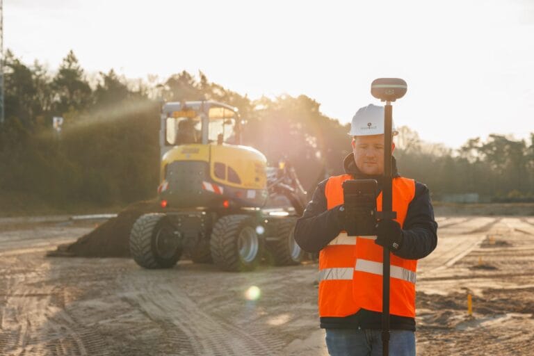 Surveyor with a UNI-GR2 pole and tablet in backlight with sun flare, yellow wheeled excavator in the background