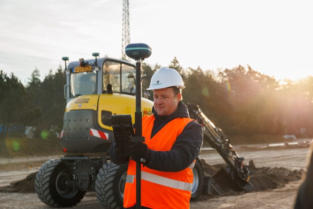 Surveyor in a white hard hat holds a UNI-GR2 pole and tablet at sunset, wheeled excavator behind