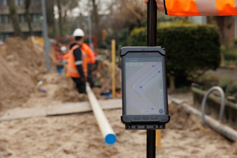 Tablet with map on a survey pole in the foreground, workers in orange hi-vis at a trench behind