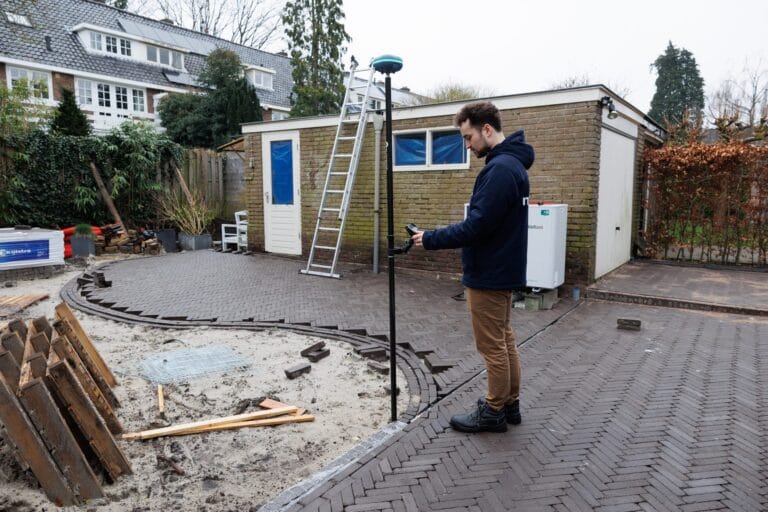 Surveyor measures a paved path with UNI-GR2 and tablet in a backyard beside a shed