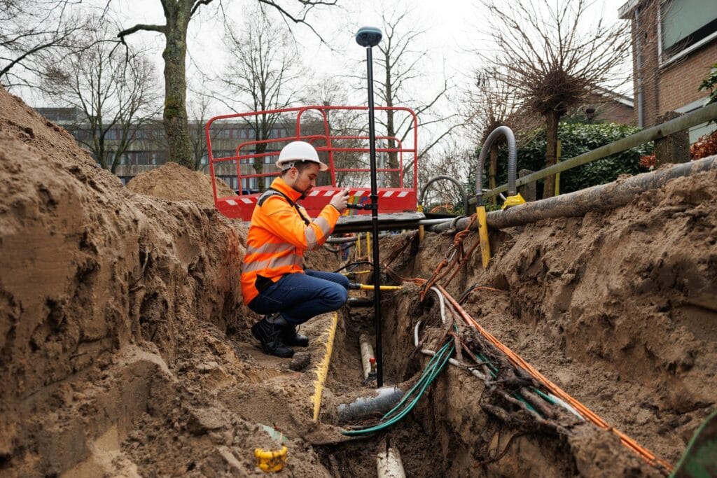 Worker measuring with a UNI-GR2 pole in a trench full of cables and pipes, red barrier and houses behind