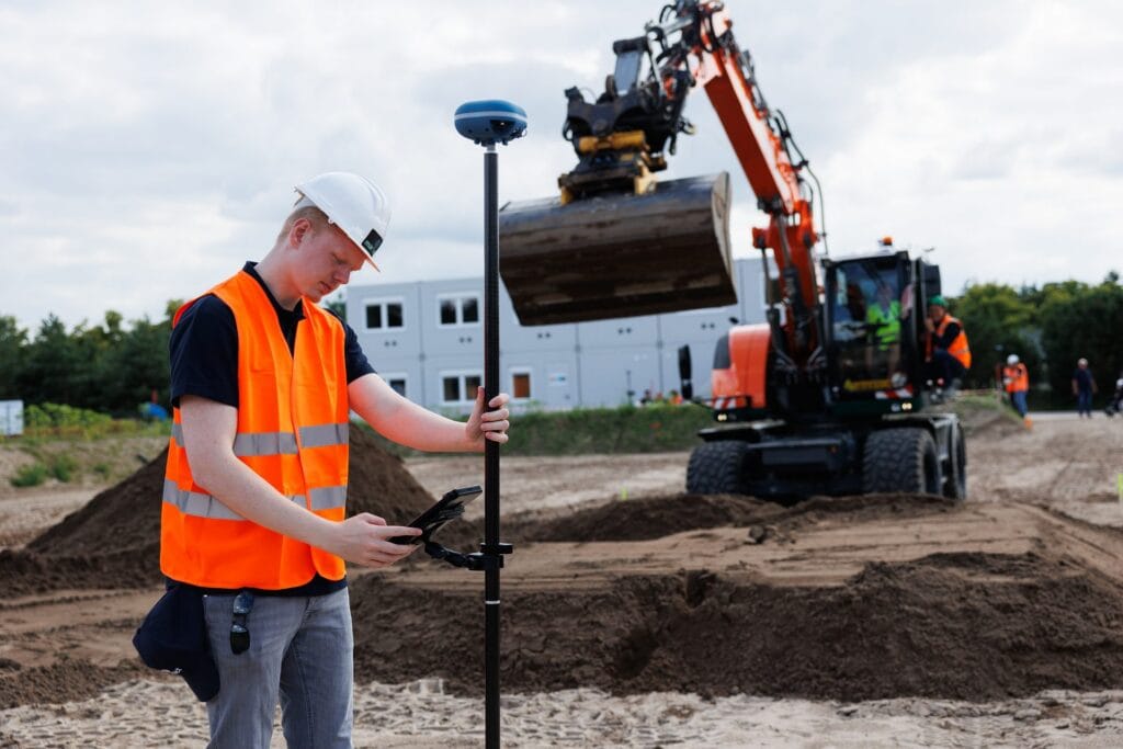 Participant with a UNI-GR2 pole checks the tablet, wheeled excavator with raised bucket behind