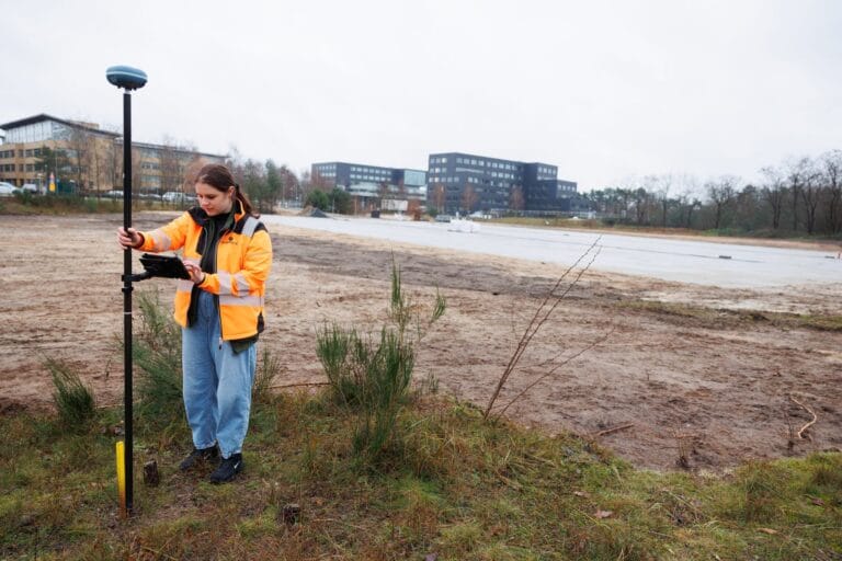 Surveyor with a UNI-GR2 rover and controller on a sand plot, office buildings behind