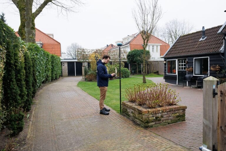 Man in a dark marXact jacket measuring with a UNI-GR2 pole and phone on a paved garden path beside hedges