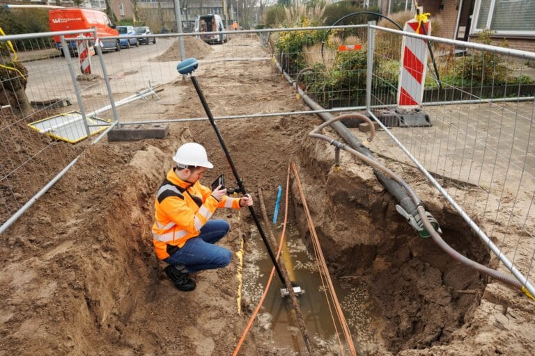 Worker in white helmet crouches in a trench with UNI-GR2 pole and tablet, site fencing and street behind