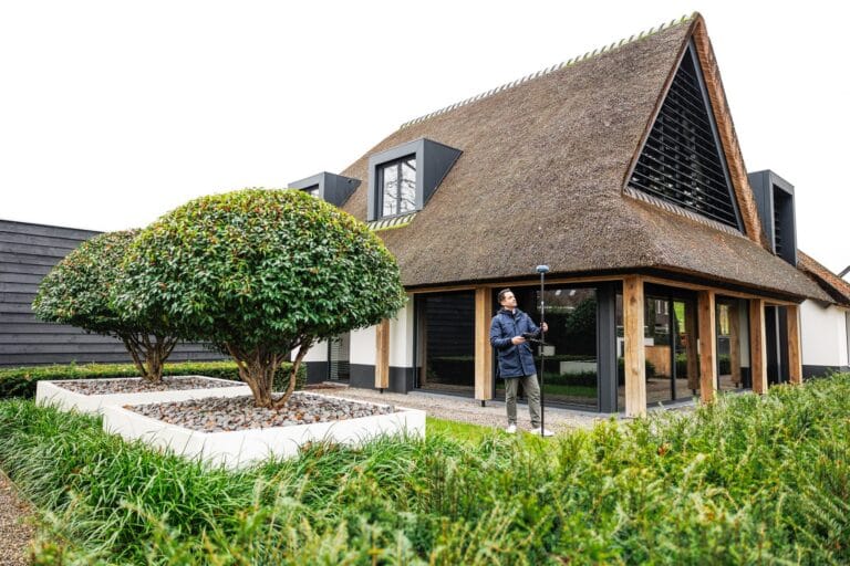 Modern villa with a thatched roof and neatly landscaped garden, a man with a GNSS pole in the background