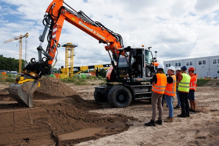 Orange wheeled excavator with machine control digging sand while visitors in hi-vis watch