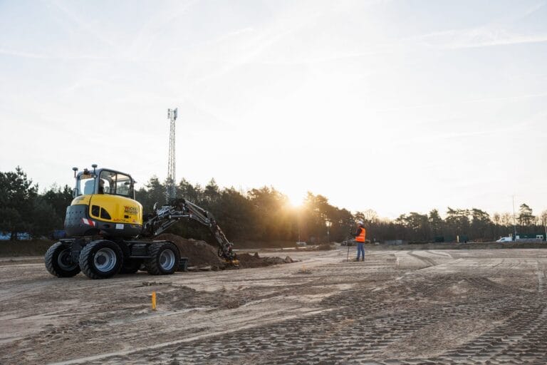 Wheeled excavator digging at sunset, yellow marker peg in the foreground and surveyor further back