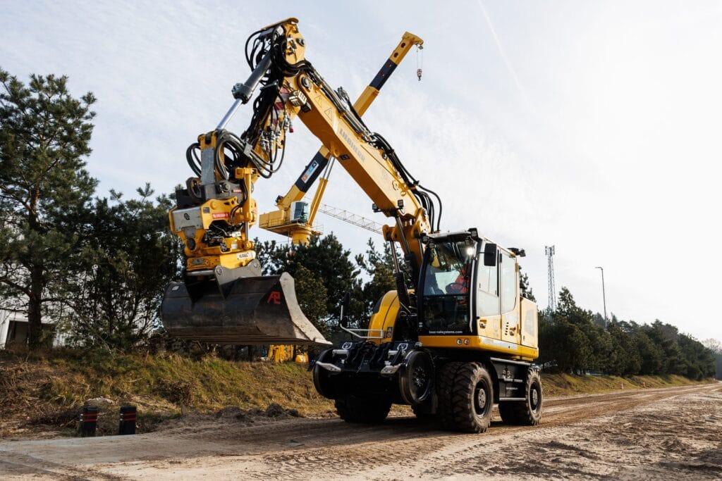 Yellow road-rail wheeled excavator with raised bucket on a sandy track between pine trees